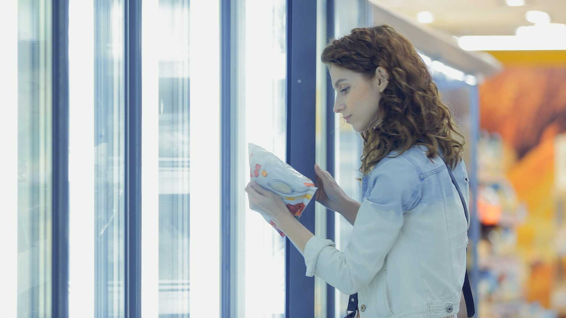 a woman looks at a food package in the frozen foods section of a grocery store